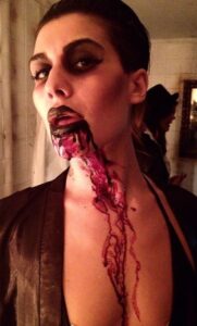 A vampy woman with Halloween makeup and vampire teeth, blood running down her neck.