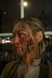 A woman with blonde hair and special effects makeup showing facial injuries.