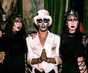 A man in a white suit with skull makeup flanked by two ladies in black with gothic makeup.