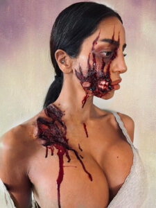 A well-endowed woman with facial and shoulder makeup showing wounds and a ripped cheek.