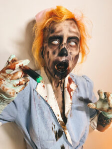 A zombie nurse with orange hair and latex gloves waves a needle.